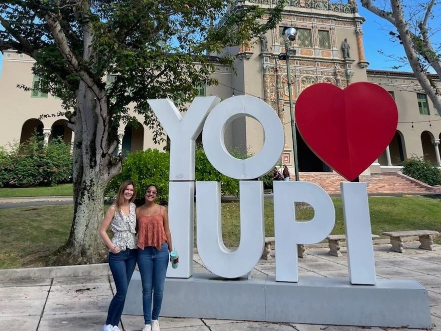 Two individuals posing in front of a large "YO ❤️ IUPI" sign