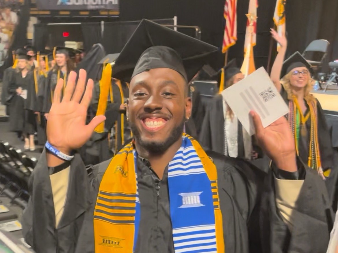 A UCF chemistry graduate in cap and gown, beaming with joy, waves to the camera at a commencement ceremony.