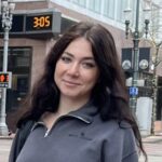 A woman with long dark hair wearing a gray zip-up jacket stands outdoors in an urban area.