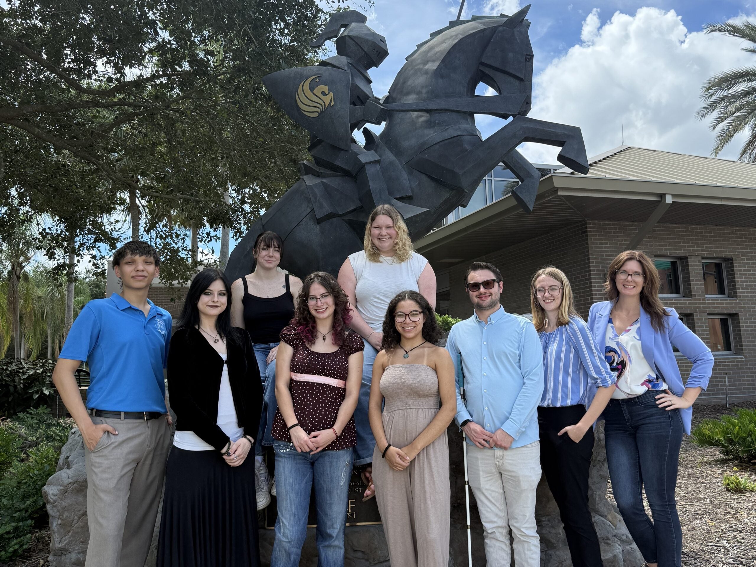 Ten people pose in front of a knight statue outdoors, with trees, a building, and a partly cloudy sky in the background. This group is part of the UCF chemistry education research team.