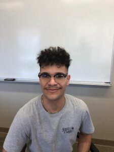 Person with curly hair and glasses smiling, seated in front of a whiteboard.
