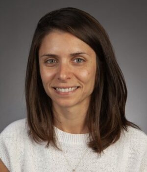 Woman with straight brown hair, wearing a white knit top, smiles at the camera against a plain gray background.