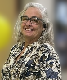An older woman with gray hair and glasses smiles while wearing a patterned blouse, standing in front of a blurred background.