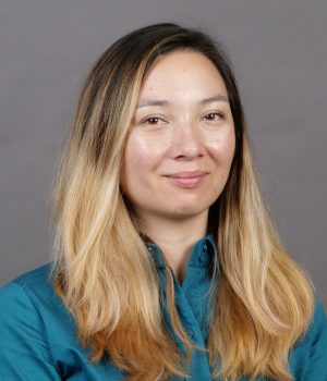 A person with long, blonde hair wearing a teal shirt is standing against a gray background.