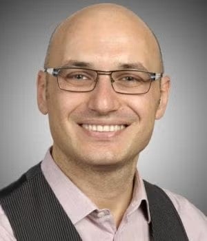 A person wearing glasses and a vest, smiling against a gray background.