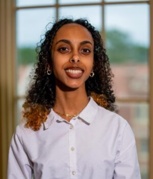 A person with curly hair wearing a white collared shirt and gold jewelry stands indoors in front of a window with a blurred outdoor background.
