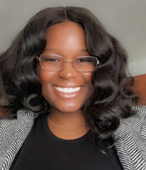 A person with wavy dark hair, wearing glasses and a striped blazer, is smiling at the camera.
