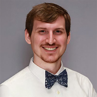A man with short brown hair, a trimmed beard, and a mustache smiles, wearing a white shirt and a navy blue bow tie with white mathematical symbols on a plain gray background.