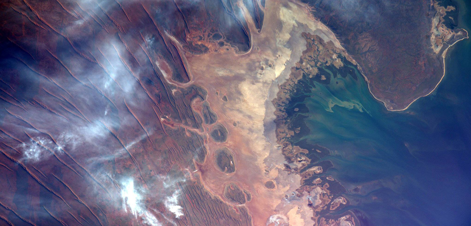 Satellite image of an arid, desert-like region with sandy textures transitioning into a coastal area with blue and green waters. Cloud streaks are visible across the landscape.