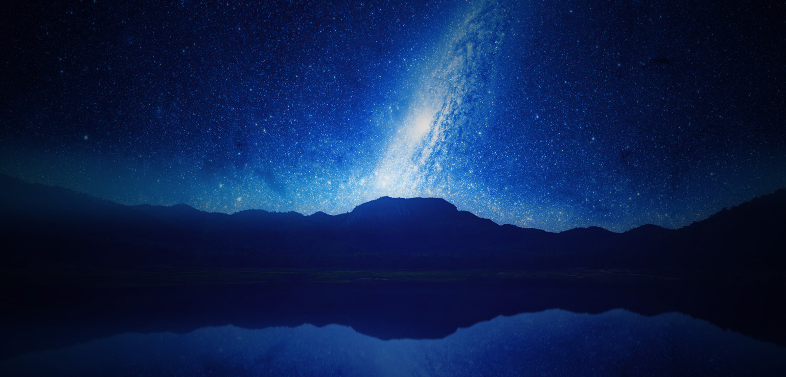 A star-filled night sky with the Milky Way visible, reflected in the still water of a lake with dark mountains in the background.