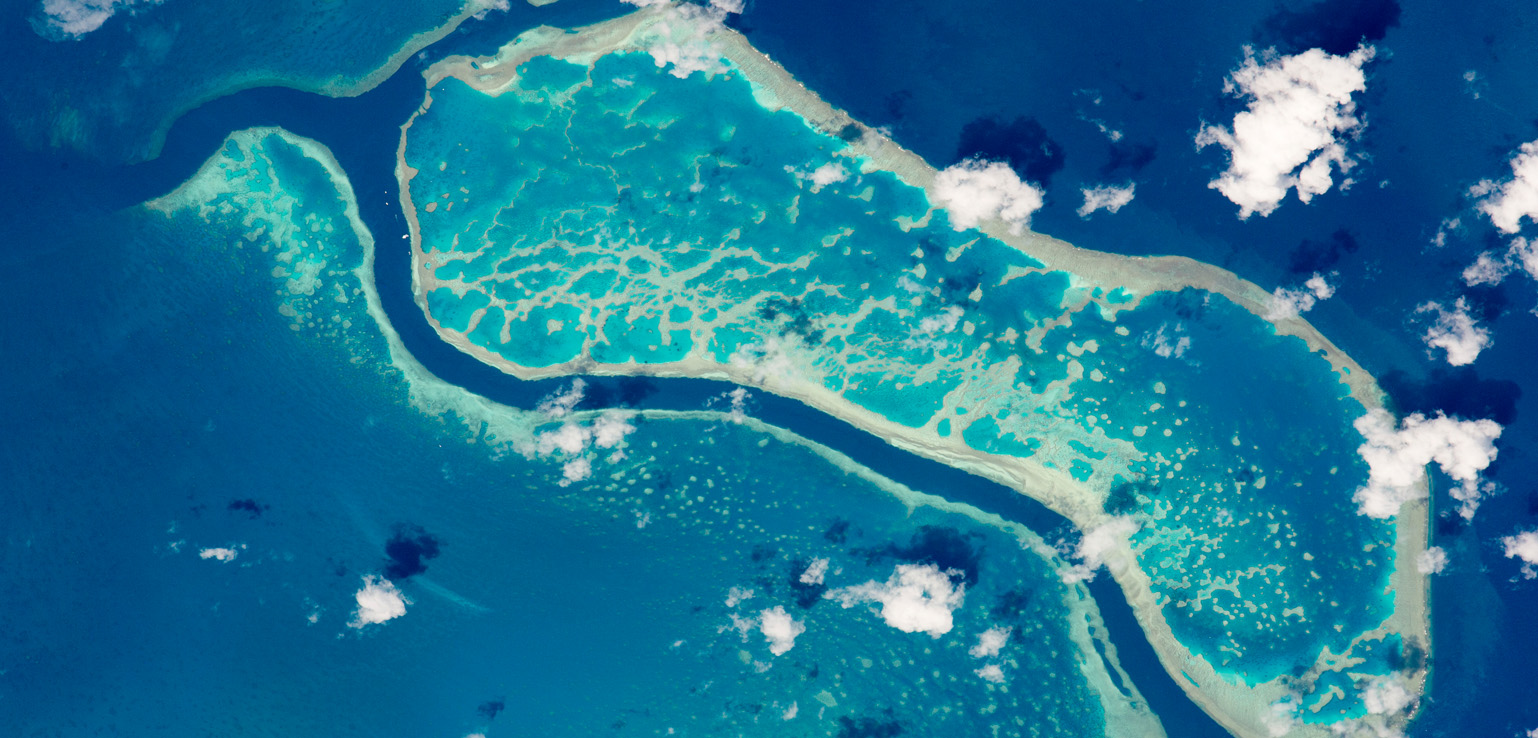 Aerial view of a coral atoll surrounded by deep blue ocean, with scattered white clouds casting shadows on the turquoise water.