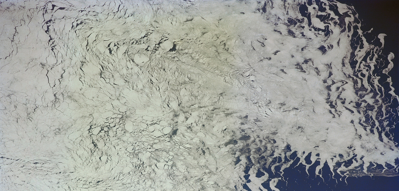 Satellite view of a vast icy terrain with fissures and icy formations extending toward the dark blue sea, showcasing the detailed texture of the ice and snow.