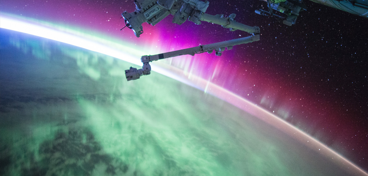 View of the aurora, with hues of green and pink, illuminating Earth’s atmosphere as seen from the International Space Station against a backdrop of stars.
