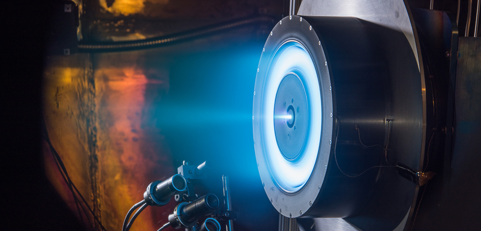 Close-up of a glowing blue electric ion thruster in operation, emitting a bright blue exhaust plume in a darkened test facility.