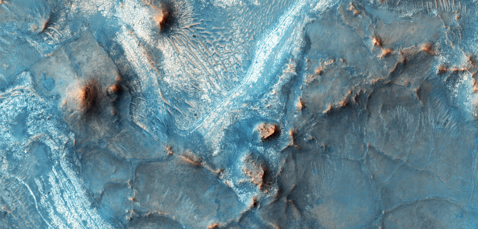 Aerial view of a Martian landscape displaying cracked patterns, elevated ridges, and scattered reddish-hued formations amidst a predominantly blue terrain.
