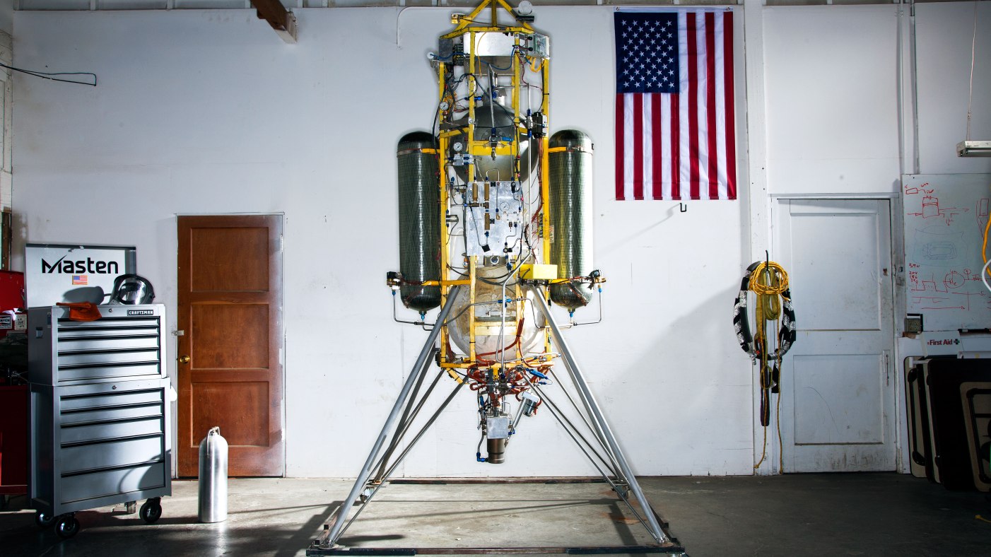A test rocket stands upright in a workshop, with an American flag hanging on the wall behind it. Nearby are various tools and equipment.