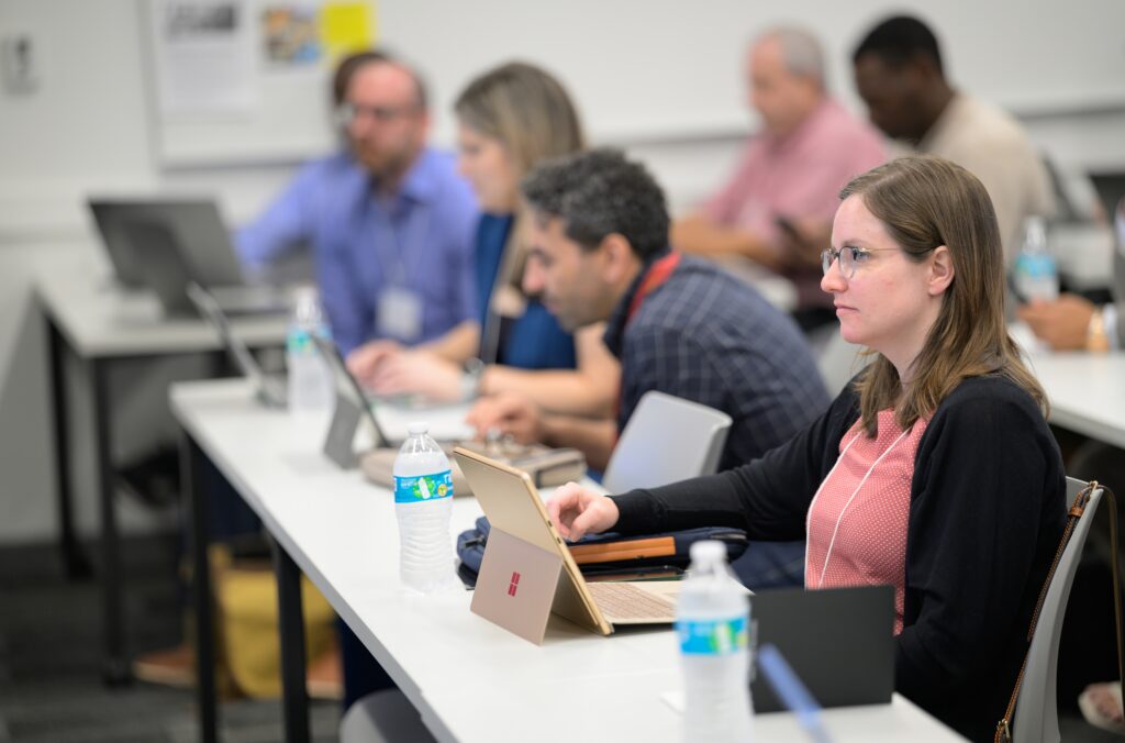 Several people sit at desks in a classroom, working on laptops and tablets, with water bottles and notebooks on the tables.