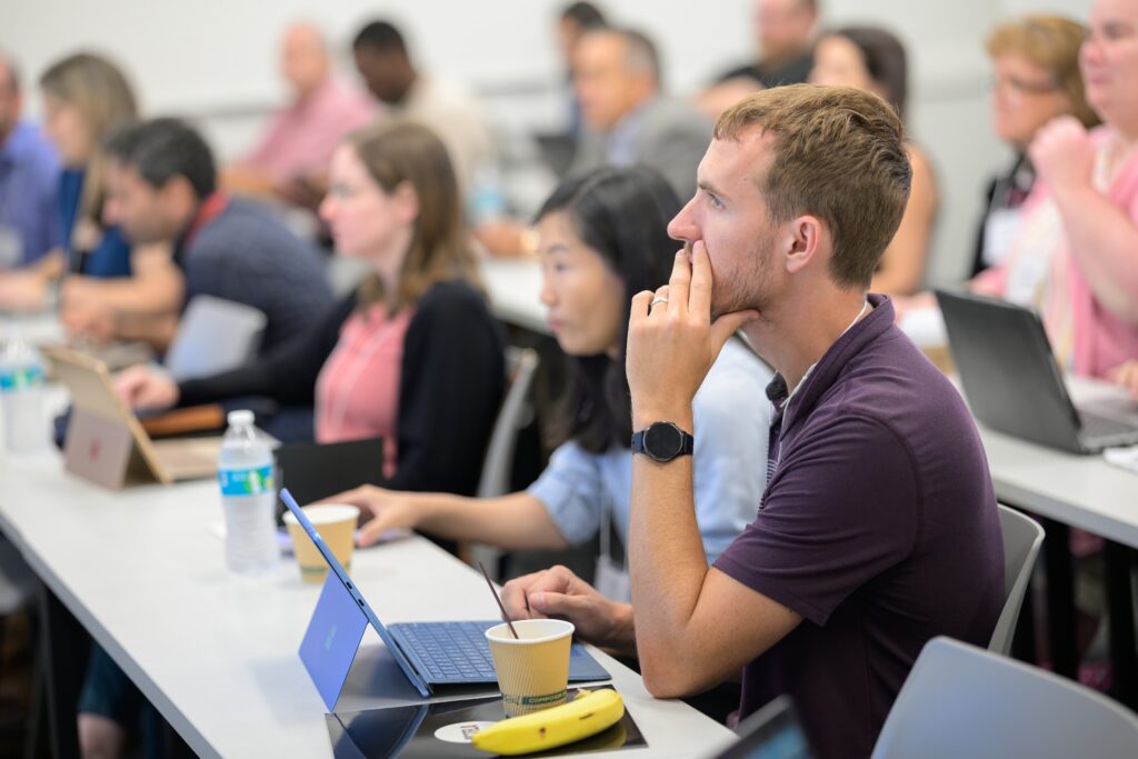 People sit at desks with laptops, notebooks, and drinks, attentively listening in a classroom or seminar setting.