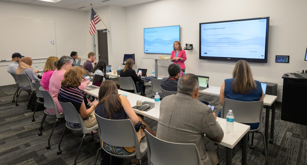 A woman stands at the front of a classroom presenting to a group of adults seated at tables with laptops and notebooks. Two large screens display presentation slides.