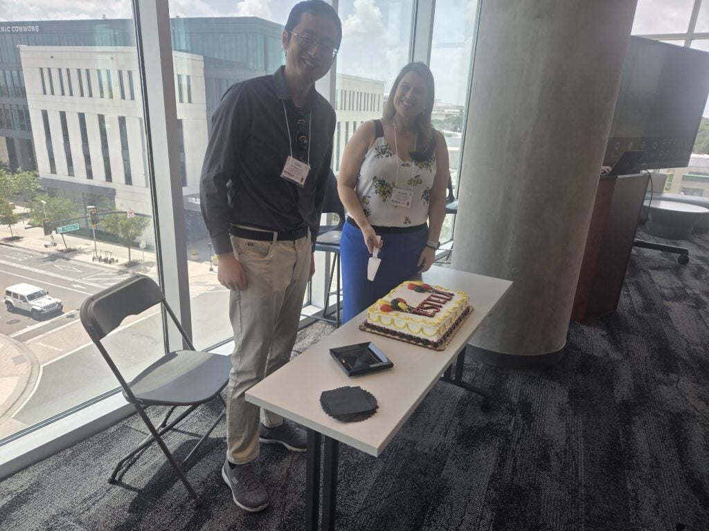 Dr. Chen and Dr. Norbutus stand by a table with a decorated sheet cake in a modern office space with large windows overlooking a city street.