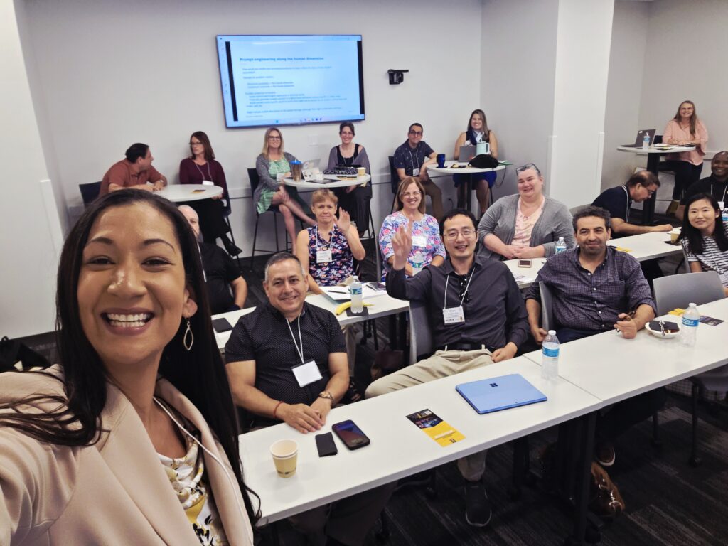 A group of people seated at tables in a classroom, some smiling at the camera; a woman in front takes a selfie. A presentation slide is displayed on a screen at the front.