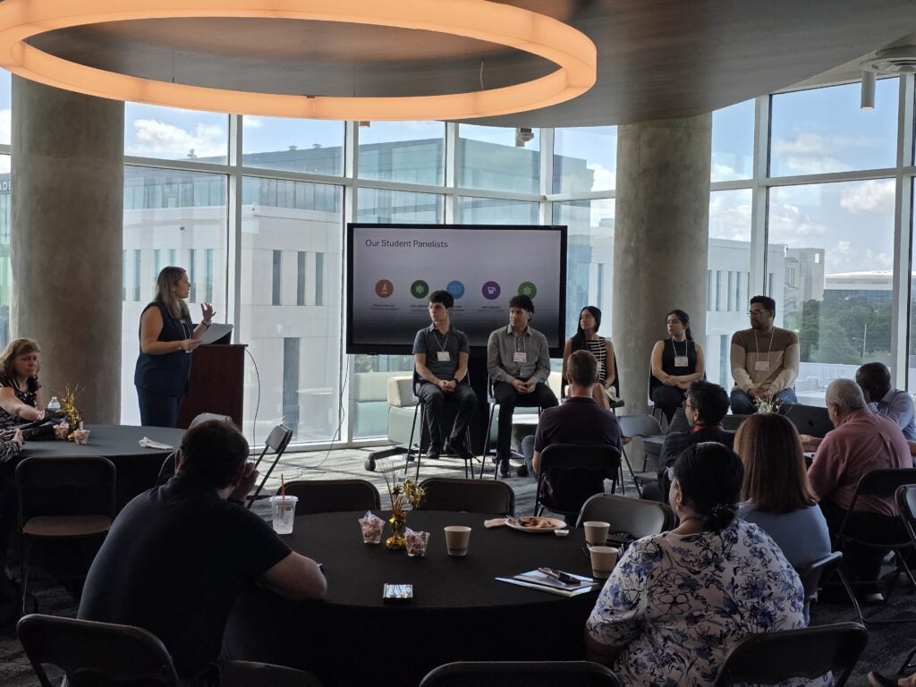 A woman speaks at a podium to an audience while six panelists sit on stage in front of a presentation screen in a modern, sunlit room.