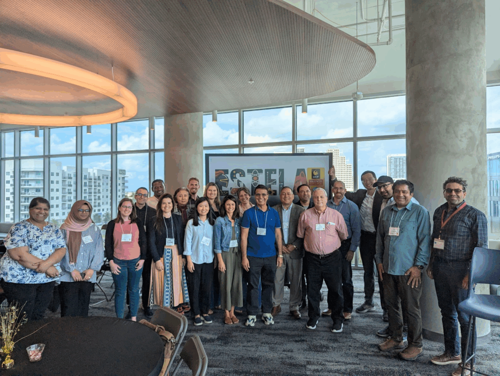 A group of ESTELA participants pose for a photo in a modern, well-lit room with large windows and city buildings visible in the background.