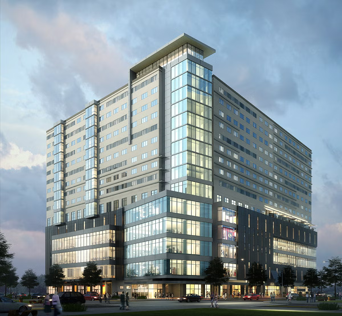 Modern multi-story office building with glass facades, illuminated at dusk, with cars and people near the entrance and trees surrounding the area.