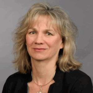 A woman with shoulder-length blonde hair wearing a black blazer, grey blouse, and a necklace, poses in front of a plain grey background.