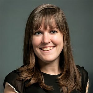 A woman with straight brown hair and bangs smiles at the camera, wearing a black top, against a plain gray background.