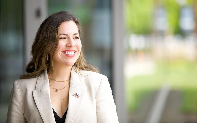 A woman wearing a light-colored blazer and necklace smiles while standing indoors near a window, with greenery visible outside.