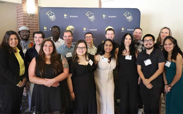 A group of people stand together indoors, posing for a photo in front of a UCF Alumni backdrop.