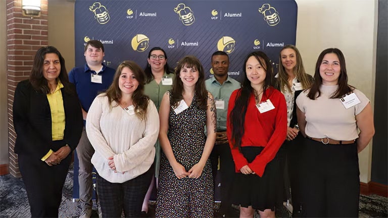 A group of nine people pose indoors in front of a UCF Alumni backdrop, wearing business casual attire and name badges.