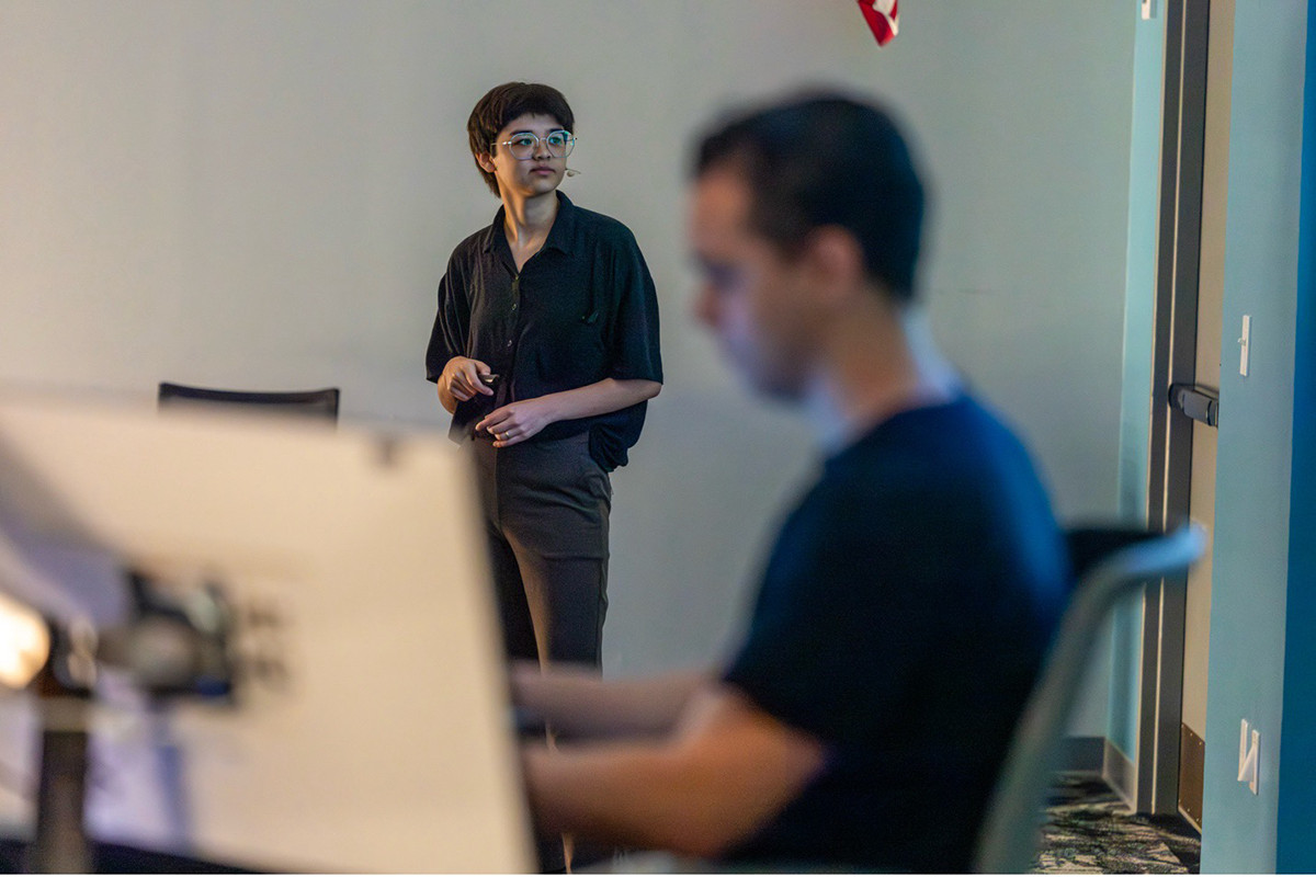 A person stands near a table, looking forward, while another individual sits in the foreground, slightly out of focus, working at a desk.