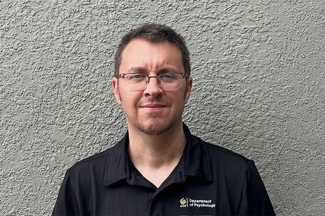A man wearing glasses and a black collared shirt with a "Department of Psychology" logo stands in front of a textured gray wall.