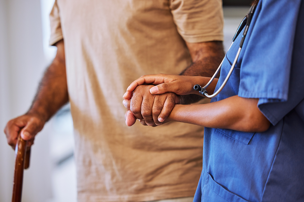 A healthcare worker in scrubs gently holds the hand of an elderly person using a cane, providing support and care.