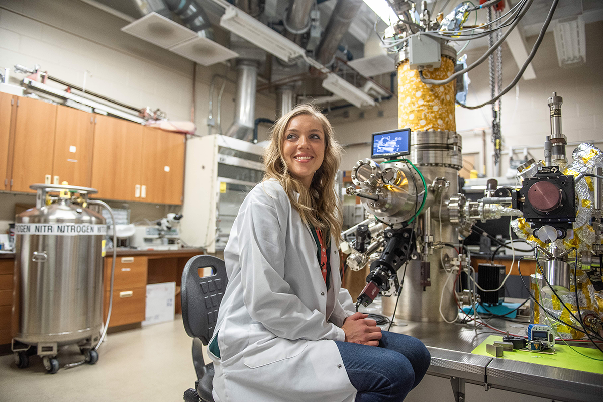 A woman in a lab coat sits at a workstation with scientific equipment in a laboratory setting, with tanks and machinery in the background.