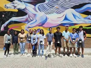 A group of twelve people stand in front of a vibrant mural featuring a winged horse. They are wearing name badges and casual clothing, posing for a group photo on a sunny day.