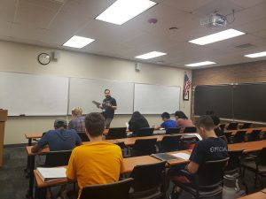 A teacher stands at the front of a classroom holding a piece of paper, instructing a group of students seated at desks facing him. The classroom has whiteboards, a clock, and an American flag on the wall.