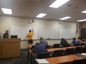 A classroom with a teacher at a podium on the left and a student in a yellow shirt writing on a whiteboard. Several students sit at desks facing the front, taking notes or reading.