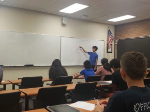 A person in a blue shirt stands at a whiteboard, explaining math to a classroom of students seated at desks. An American flag hangs on the brick wall next to the whiteboard.