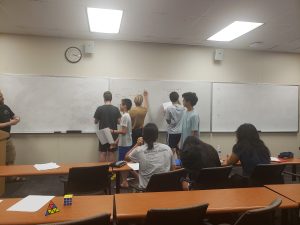 Students are standing and writing on a large whiteboard in a classroom while others are seated, watching or taking notes.