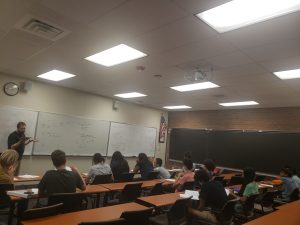 A classroom with students seated at desks facing a teacher who is writing on a whiteboard filled with mathematical equations. An American flag hangs on the wall, and there are fluorescent lights on the ceiling.