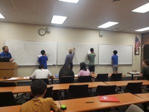 Four individuals writing on a large whiteboard in a classroom. Others are seated at desks, observing. An instructor stands by a podium on the left, and an American flag is displayed in the background.