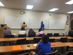 A classroom scene with a man writing on a whiteboard while another person, standing nearby, looks on. Several students sit at desks, facing the board and engaged in the lecture. An American flag is visible on the wall.