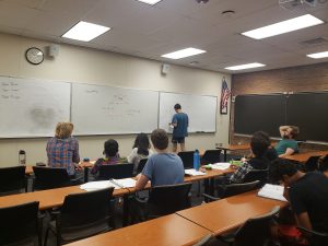 Students in a classroom sitting at desks and facing a whiteboard with mathematical problems written on it. One student is standing at the front, solving a problem on the board. An American flag is on the right wall.