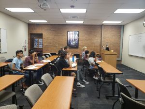 A group of students sits attentively at desks in a classroom facing a teacher who is at a lectern. Tables and chairs are arranged in rows. A painting and whiteboard are visible on the walls.