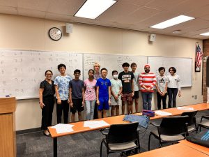 A group of twelve people stands in front of a large whiteboard in a classroom. There are papers and chairs in the foreground, and a clock and an American flag on the wall.