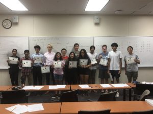 A group of ten people standing in a classroom, each holding a certificate. They are smiling at the camera, and the background shows a whiteboard with various notes and equations.