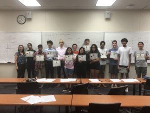 A group of students and two adults are standing in a classroom holding certificates. A whiteboard with writing is in the background.